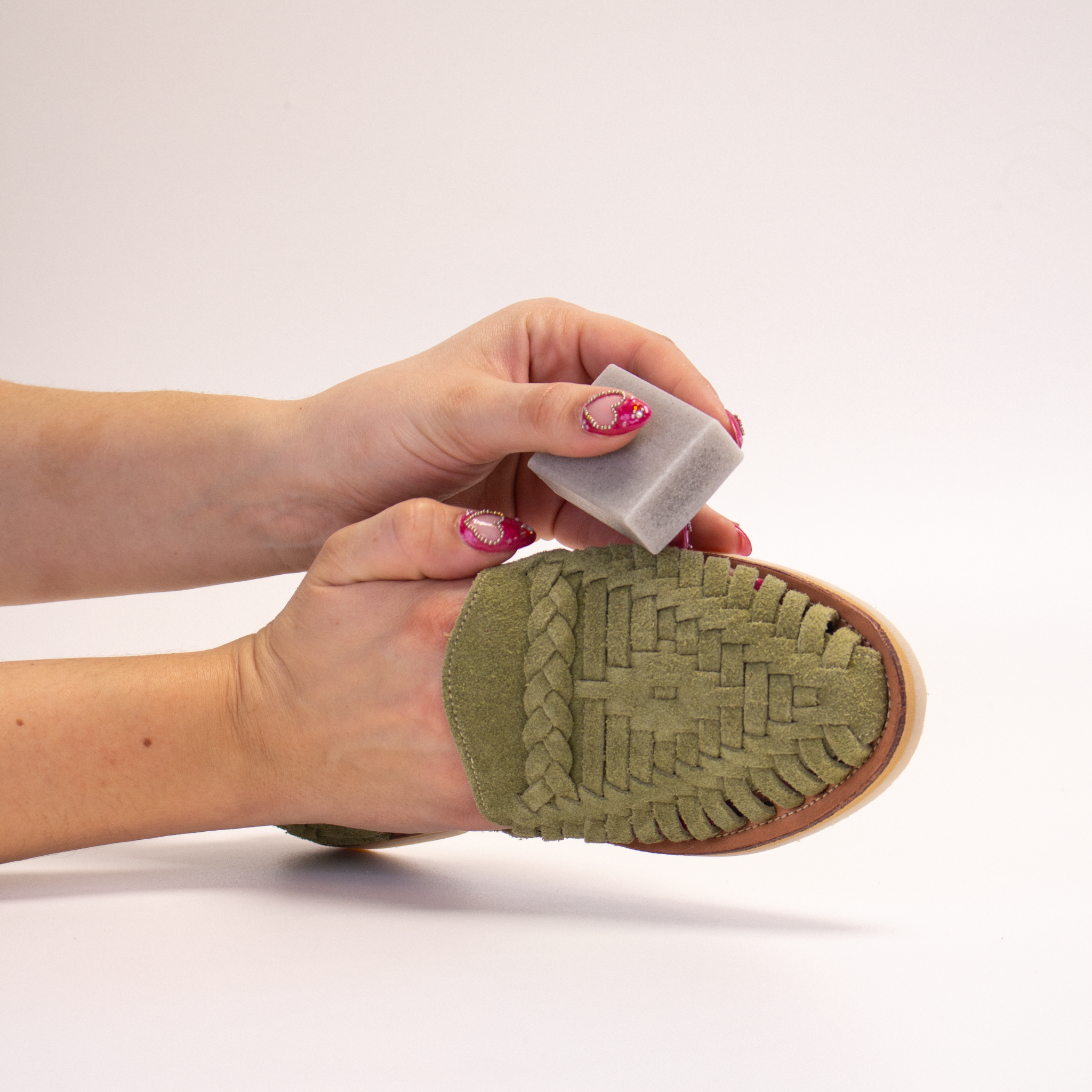 Une main maintient la huarache Alegre Suède Olive pour mettre en valeur son tressage, tandis que l’autre frotte délicatement la GOMME NET de FAMACO sur le dessus de la chaussure.