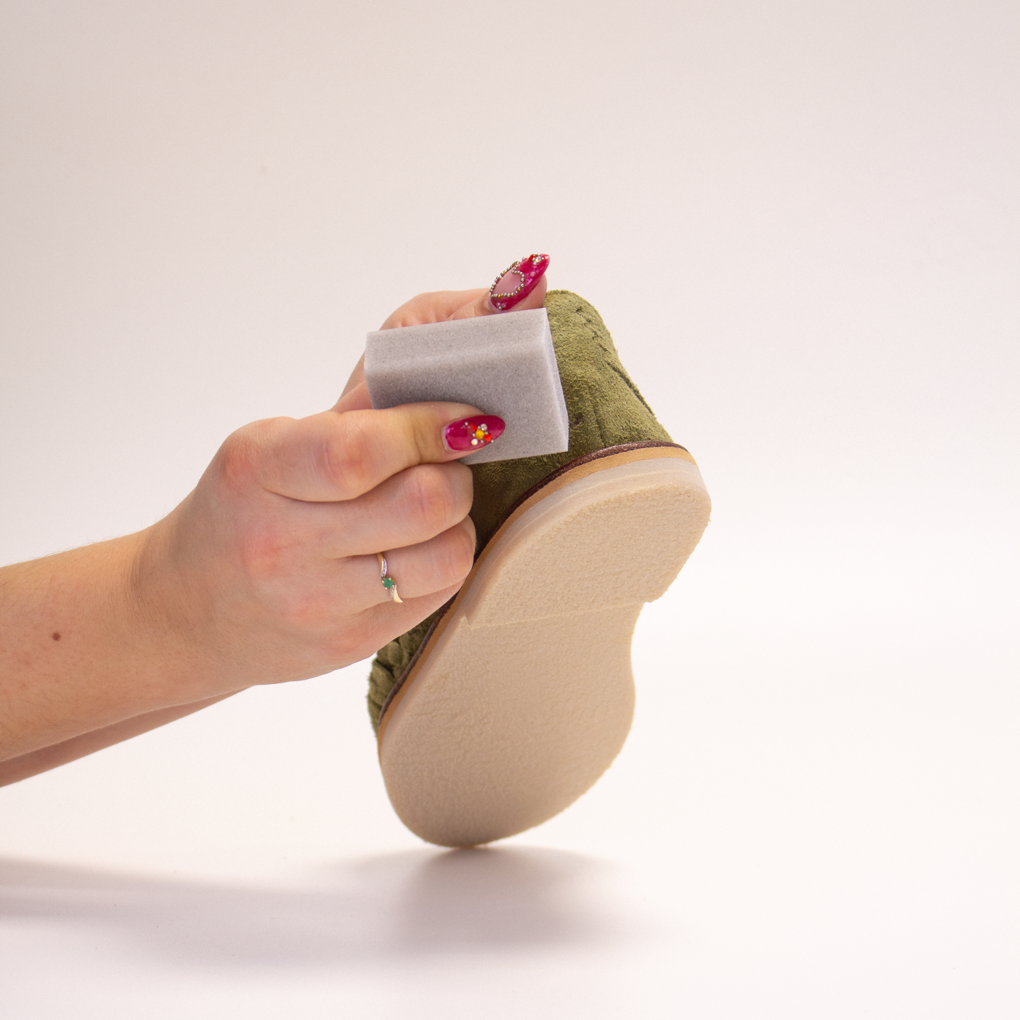 Une main tient la huarache Alegre Suède Olive en mettant en avant l’arrière, tandis que l’autre frotte la GOMME NET de FAMACO pour nettoyer la chaussure.