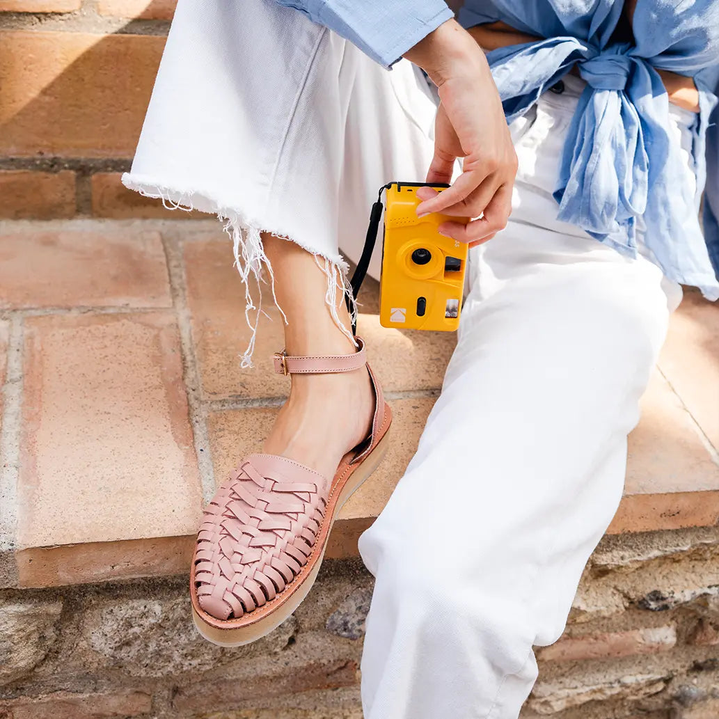 Une femme assise sur un mur en brique relève un pied pour montrer ses Sandales Loma Blush. Elle tient un appareil photo jetable entre les mains.