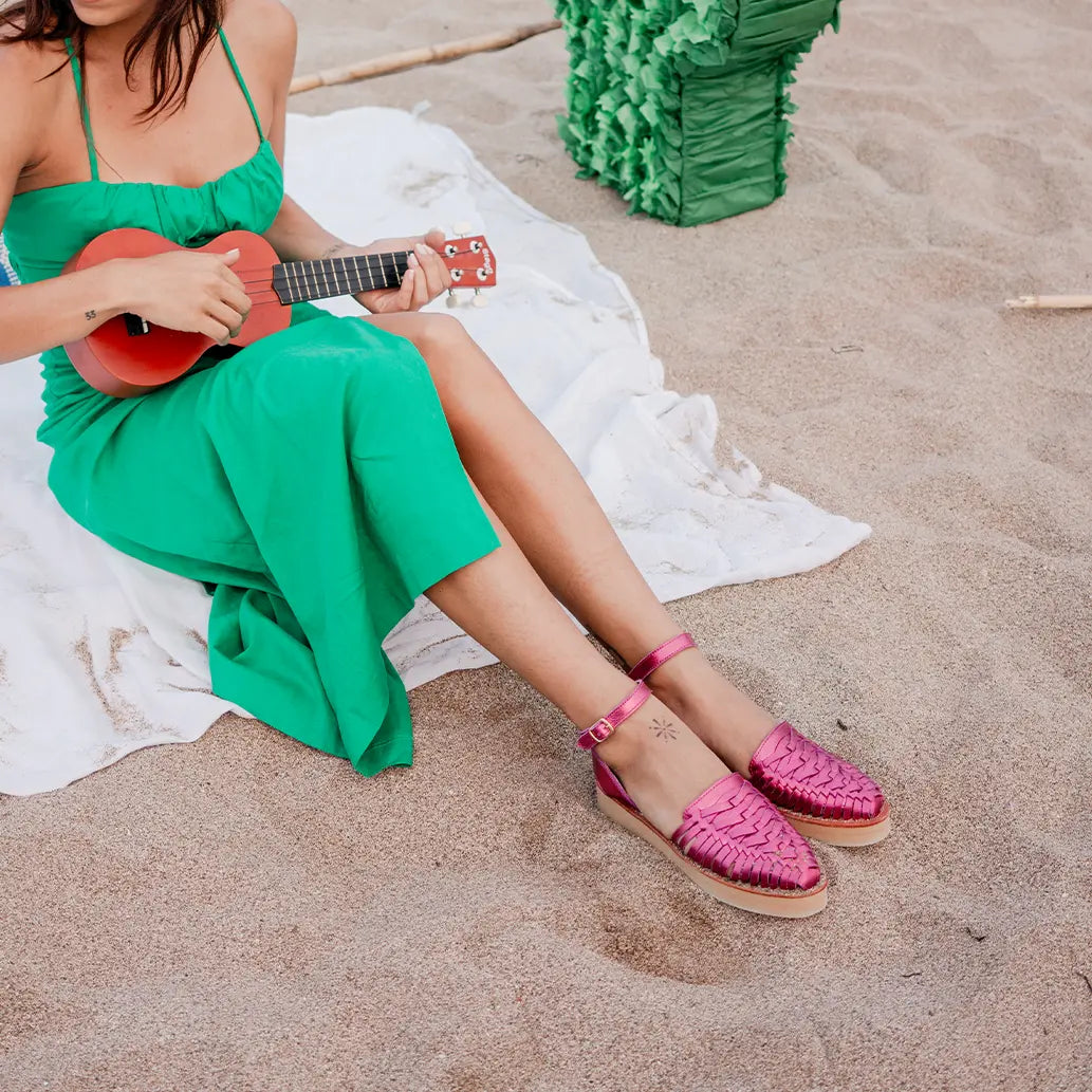 Une femme jouant du Ukulélé sur la plage porte les Sandales Loma Rose Flashy ainsi qu'une longue robe verte.