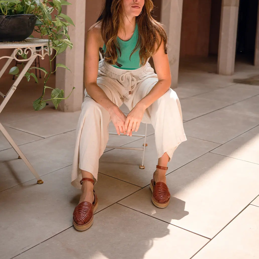 Une femme assise de manière décontractée sur une chaise de jardin en métal blanc porte les Sandales Loma Tabac.