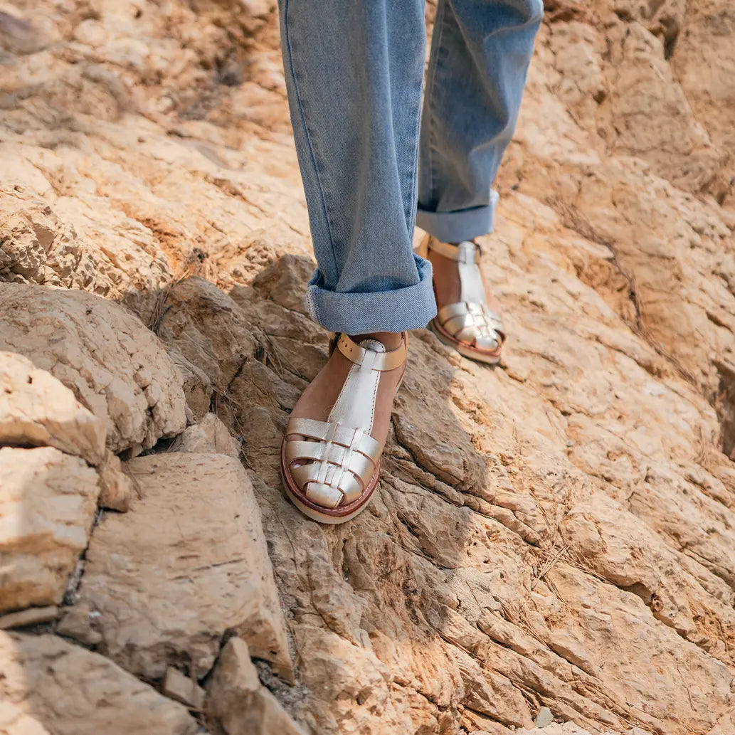 Une femme marche sur une falaise au soleil avec les Sandales Matilda Champagne.