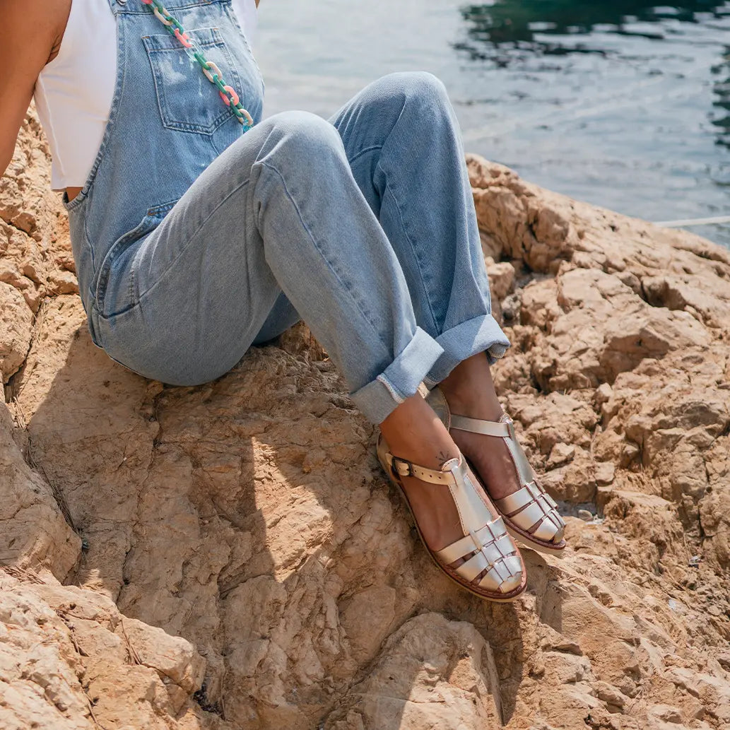 Une femme est assise sur une falaise au bord de l'eau avec les Sandales Matilda Champagne. Elle porte une salopette en jean bleu.