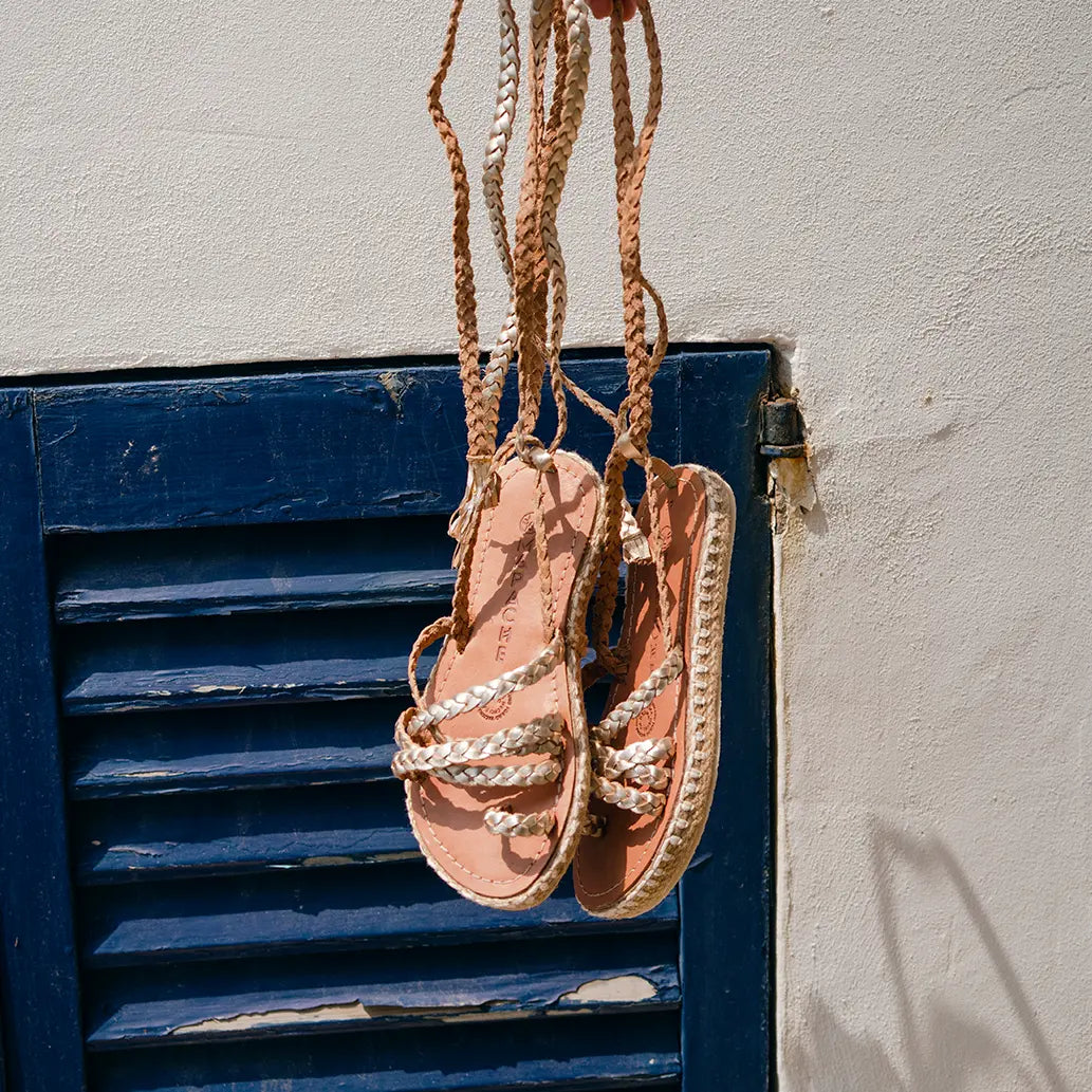 Les Sandales Ola Champagne sont suspendues devant un mur blanc avec une fenêtre bleue marine.
