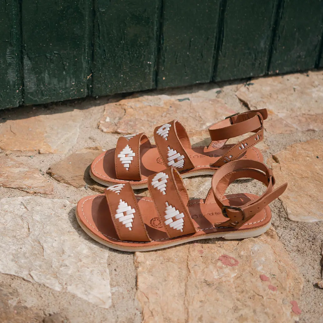 Les Sandales Rosa Camel Blanc sont posées sur de l'ardoise beige et devant une porte en bois vert sapin.