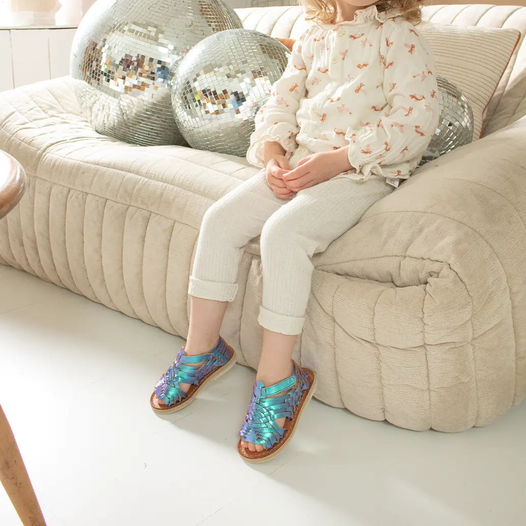 Une petite fille est assise sur un canapé beige rempli de boules à facettes et porte les Sandales Sandalita Blue Mermaid ainsi qu'un legging beige côtelé et une blouse de la même couleur.