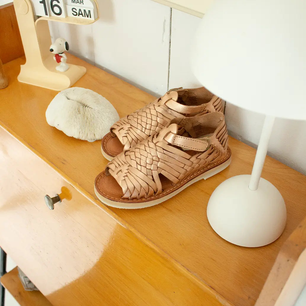 Les Sandales Sandalita Or Rose sont posées sur une étagère en bois à côté d'une lampe blanc et d'un calendrier.