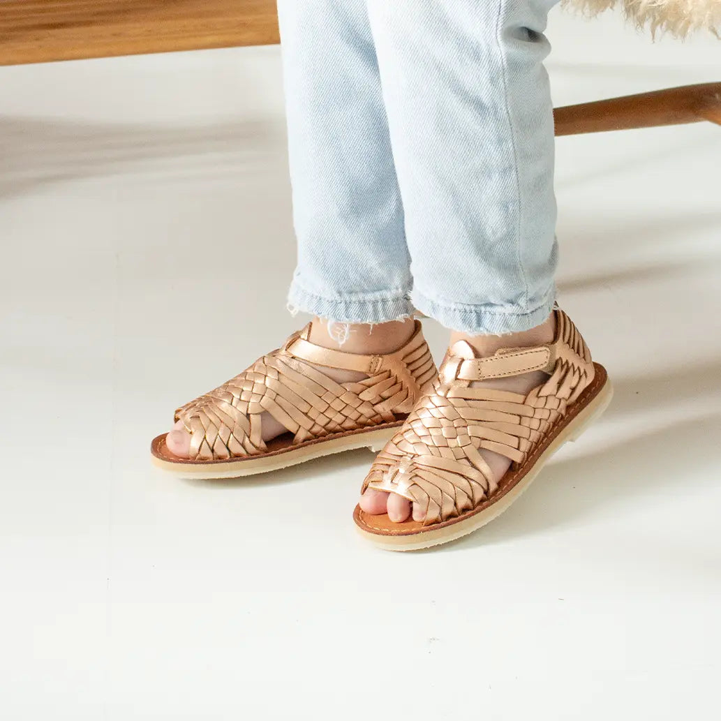 Une petite fille souriante dans un fauteuil en bois avec un coussin en poils longs camel porte les Sandales Sandalita Or Rose.