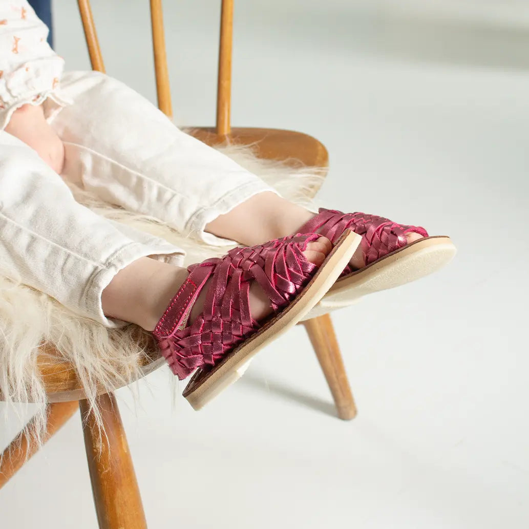 Une petite fille assise sur une chaise en bois porte les Sandales Sandalita Rose Flashy ainsi qu'un jean blanc.