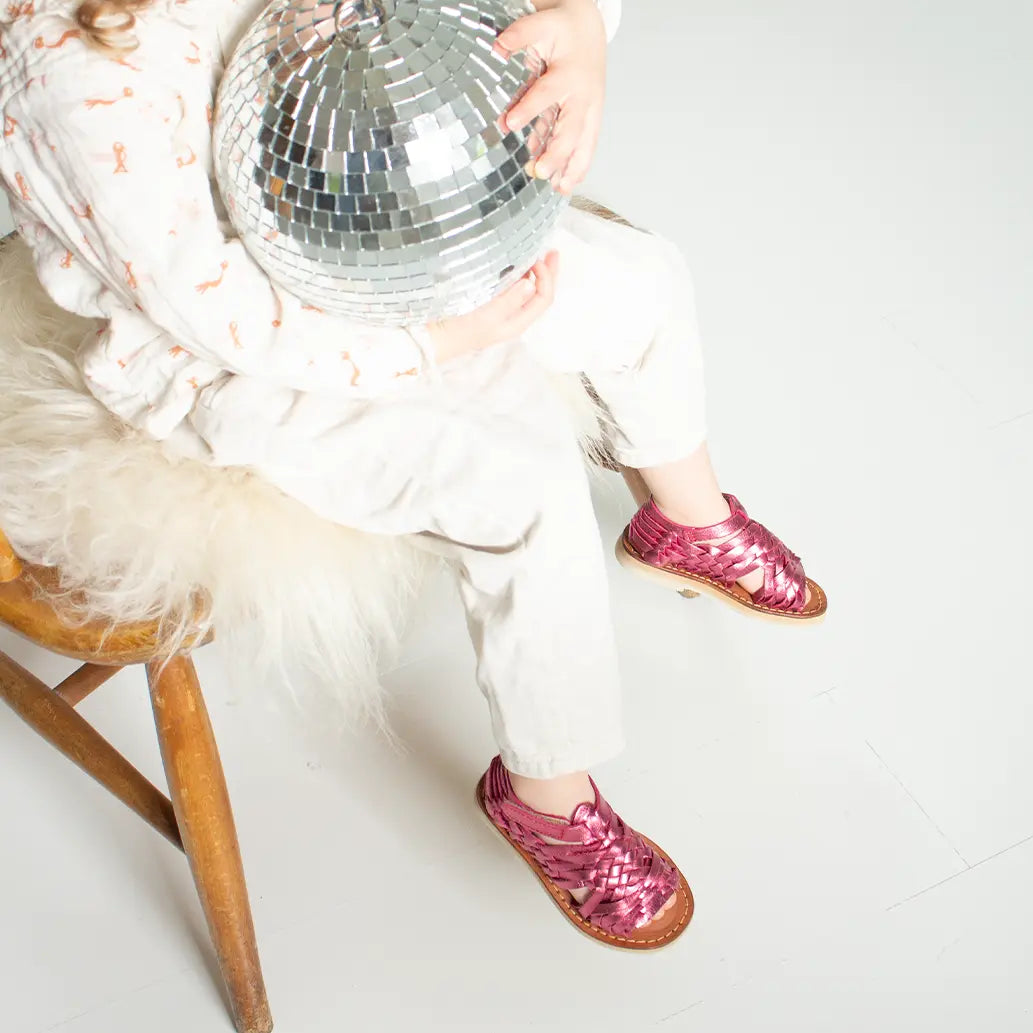 Une petite fille assise sur une chaise en bois porte les Sandales Sandalita Rose Flashy. Elle tient dans ses mains une boule à facettes et porte également un jean blanc.