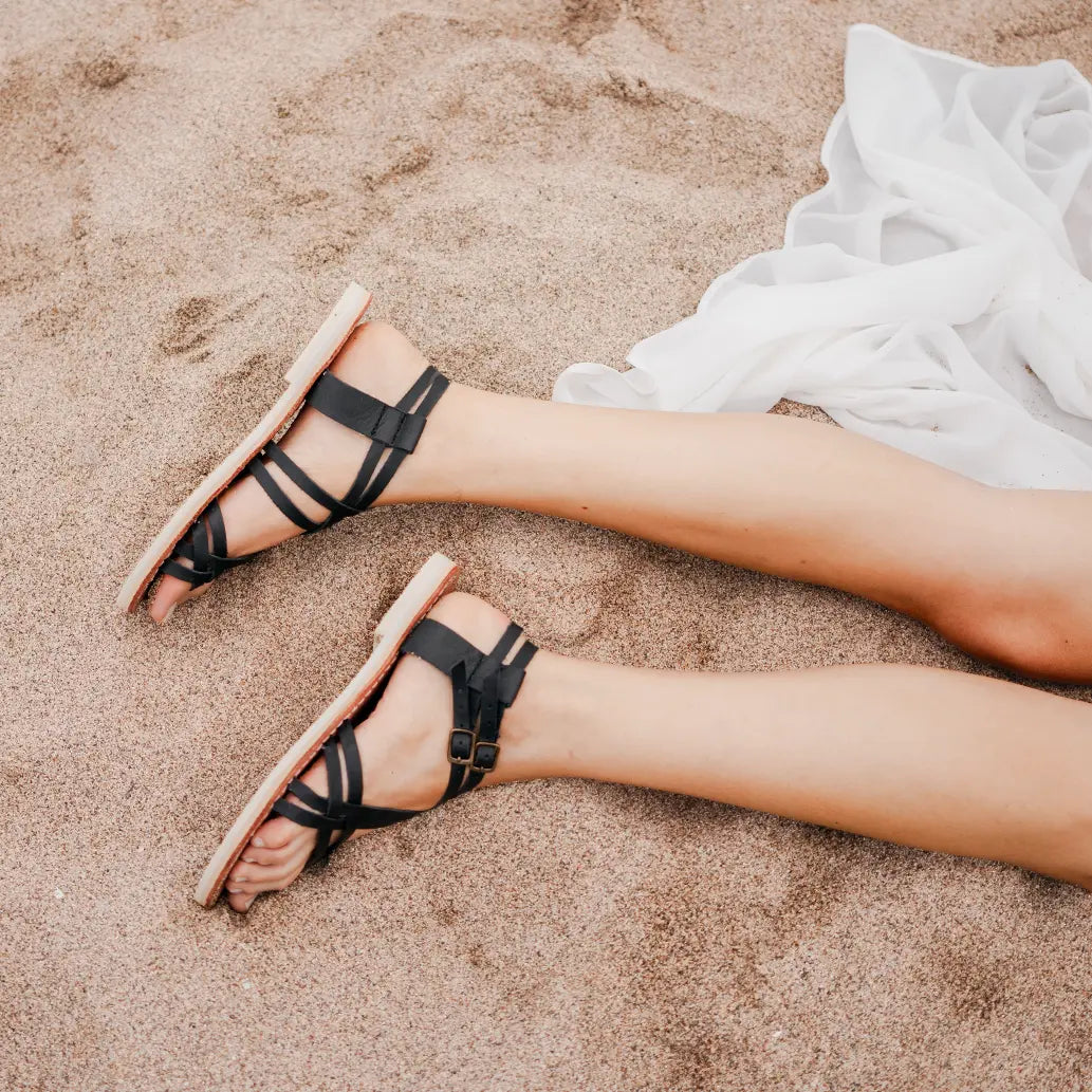 Une femme est allongée sur le côté dans le sable et porte les Sandales Selena Noir.