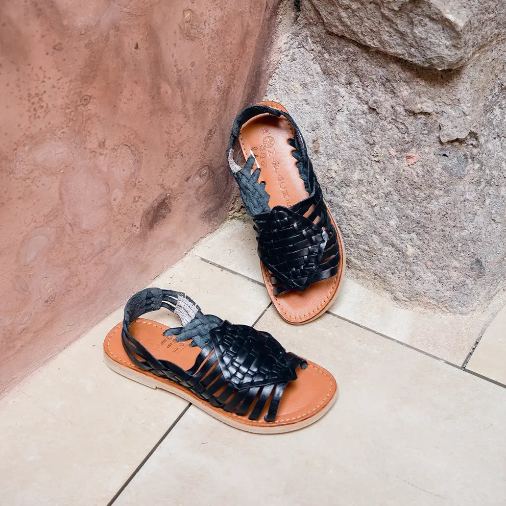 Les Sandales Leonora Noir sont appuyées contre un mur en pierre grise et reposent sur un carrelage blanc.