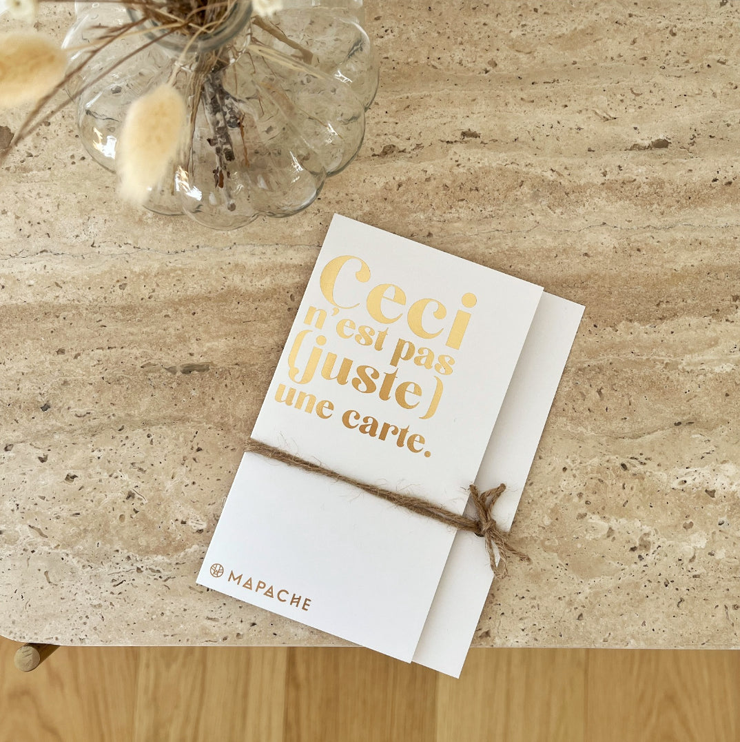 Une carte avec écrit "Ceci n'est pas une carte" repose sur commode en pierre, un vase avec des fleurs sèches à côté.