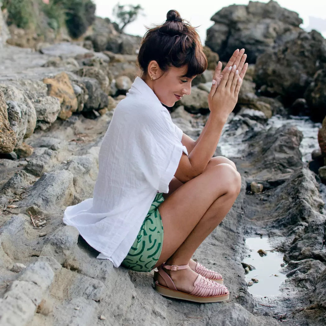 Une femme accroupie sur une crique au bord de l'eau porte les Sandales Loma Blush. Elle sourit et se frotte les mains.