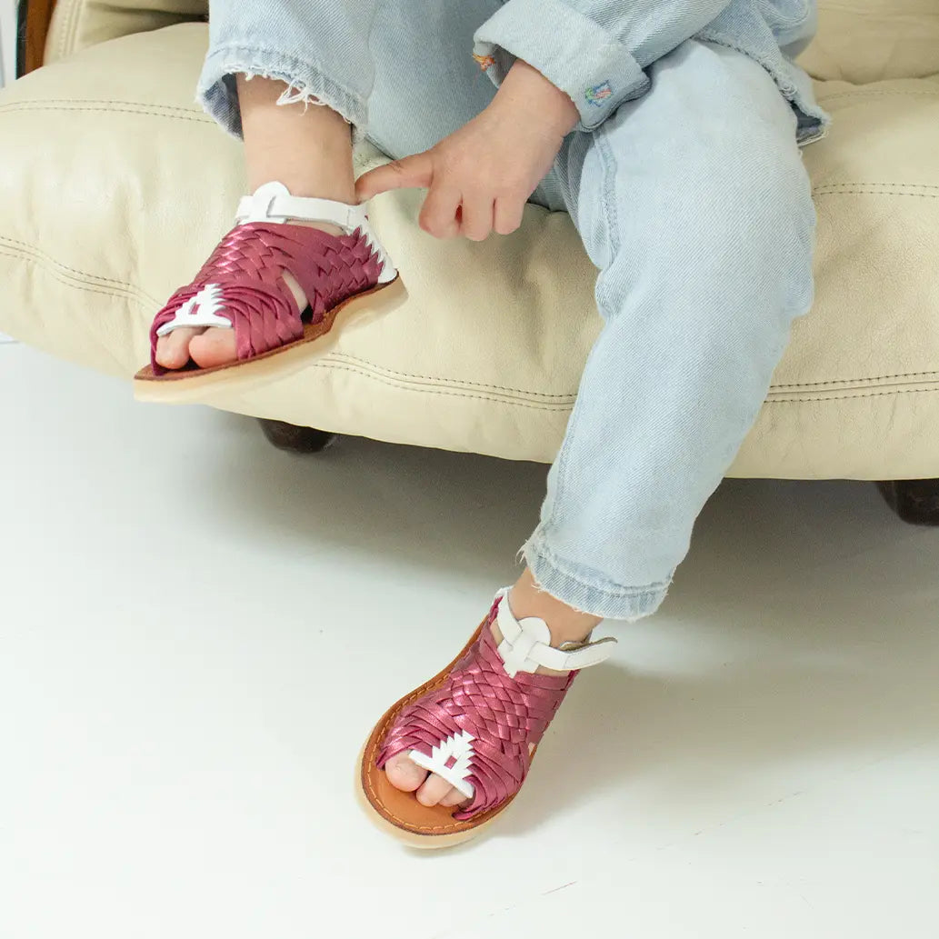 Une petite fille assise sur un fauteuil beige réajuste l'arrière de ses Sandales Sandalita Rose Flashy Blanc.