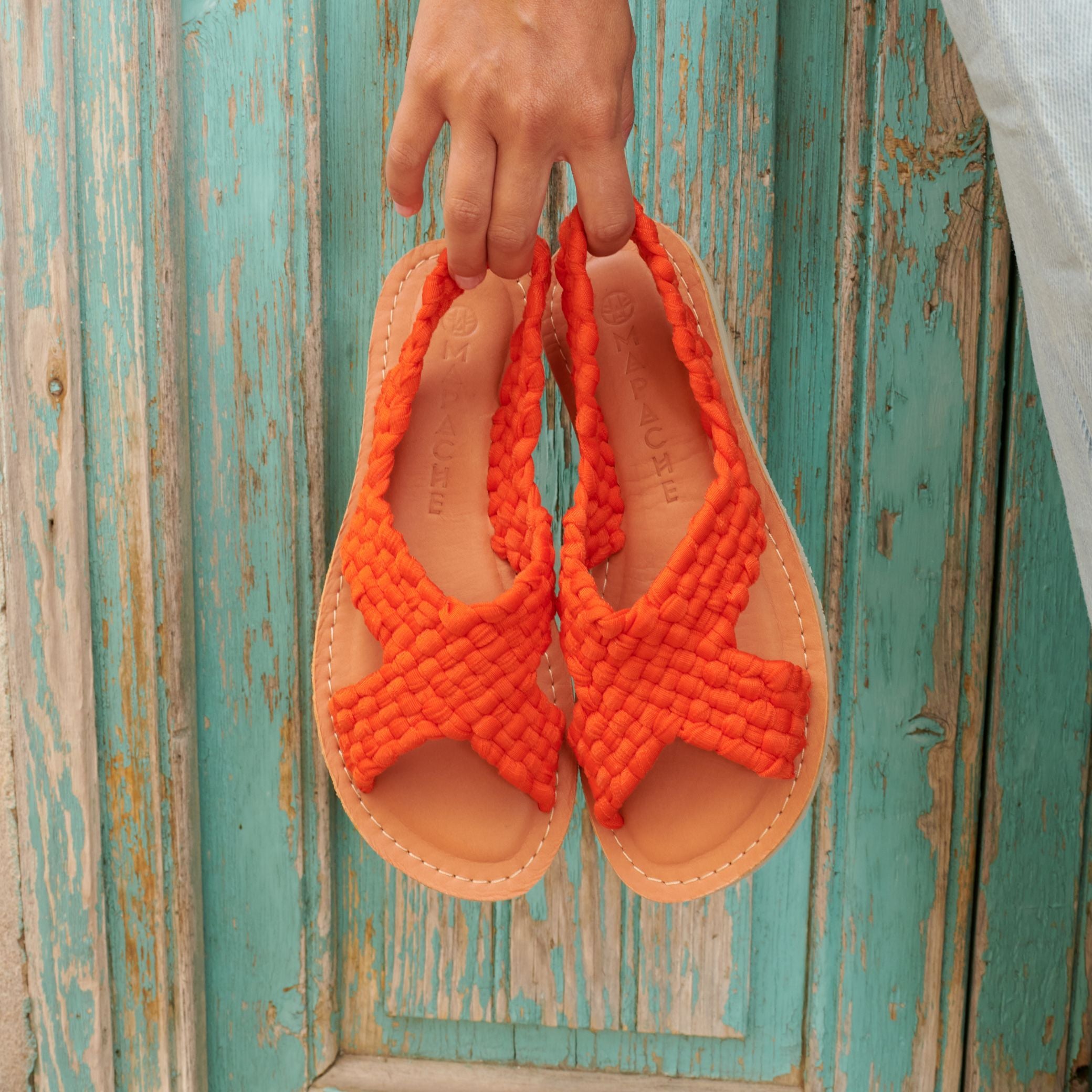 Les sandales Zula Sanguine au tressage orange, sont tenues à la main par Laura.